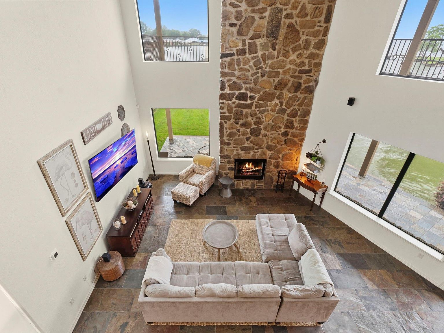 Soaring great room at 295 Bayview Lane seen from the loft balcony, a two-story stone wood-burning fireplace anchoring the open-concept living area with Lake Livingston framed by windows on three sides