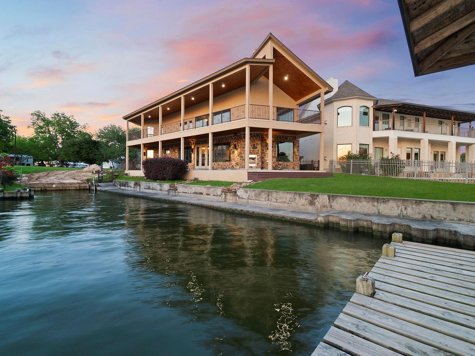 Twilight view of 295 Bayview Lane from the water, a stone-and-cream two-story lakefront residence on Lake Livingston with a covered upper balcony reflected in the canal at golden hour